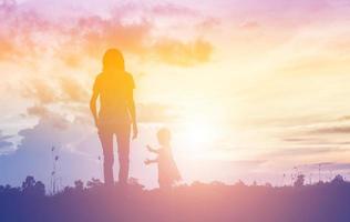 una silhouette di una bambina felice le braccia della sua amorevole madre per un abbraccio, davanti al tramonto nel cielo in una giornata estiva. foto