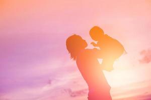 una silhouette di una bambina felice le braccia della sua amorevole madre per un abbraccio, davanti al tramonto nel cielo in una giornata estiva. foto