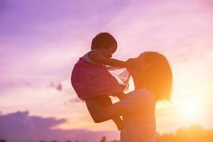 una silhouette di una bambina felice le braccia della sua amorevole madre per un abbraccio, davanti al tramonto nel cielo in una giornata estiva. foto