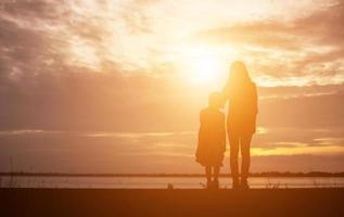 una silhouette di una bambina felice le braccia della sua amorevole madre per un abbraccio, davanti al tramonto nel cielo in una giornata estiva. foto