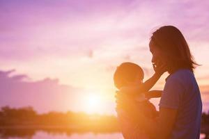 una silhouette di una bambina felice le braccia della sua amorevole madre per un abbraccio, davanti al tramonto nel cielo in una giornata estiva. foto