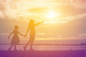 una silhouette di una bambina felice le braccia della sua amorevole madre per un abbraccio, davanti al tramonto nel cielo in una giornata estiva. foto