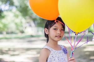 bambina sveglia che tiene palloncini colorati nel prato contro il cielo blu e nuvole, allargando le mani. foto