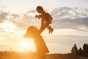 una silhouette di una bambina felice le braccia della sua amorevole madre per un abbraccio, davanti al tramonto nel cielo in una giornata estiva. foto