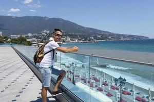 giovane uomo attraente con i capelli scuri in occhiali da sole e uno zaino su un moderno terrapieno in riva al mare in una città europea. vista laterale. foto