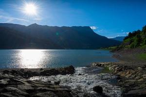 piccola cascata in una giornata di sole che entra in un grande lago con le montagne sullo sfondo foto