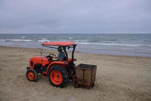 camion della spazzatura uomo lavoratore sulla spiaggia. il trattore del conducente organizza la spazzatura. una macchina speciale rossa filtra la sabbia e ne rimuove i rifiuti sulla spiaggia. macchina per la pulizia dei rifiuti da spiaggia al lavoro. foto