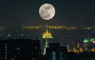 luna piena surreale sopra la pagoda dorata, punto di riferimento a bangkok, tailandia, con vista sul paesaggio urbano foto