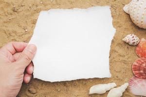 concetto mano che tiene un foglio bianco bianco su uno sfondo marrone di sabbia e conchiglia, carta bianca per la tua lettera. foto