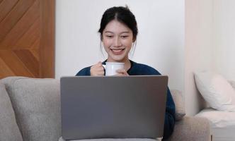 ragazza asiatica che lavora al computer portatile online, utilizzando internet e tiene la tazza. seduto sul divano di casa. foto