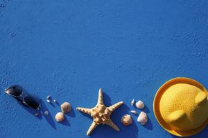 cornice con accessori da spiaggia su un cappello di paglia giallo a tema nautico, occhiali da sole, stelle marine e conchiglie su sfondo blu. concetto di vacanza, viaggio in mare, protezione dai raggi UV, nuoto. copia spazio. piatto foto