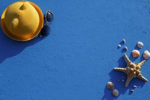 cornice con accessori da spiaggia su un cappello di paglia giallo a tema nautico, occhiali da sole, stelle marine e conchiglie su sfondo blu. concetto di vacanza, viaggio in mare, protezione dai raggi UV, nuoto. copia spazio. piatto foto