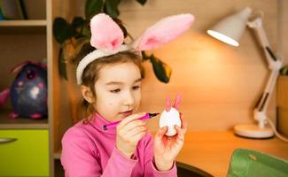 una ragazza nelle orecchie di un coniglio pasquale dipinge le uova con un pennarello all'interno della casa. artigianato, preparazione per una festa religiosa, vassoio con uova, orecchie di lepre fatte di plastilina foto