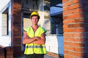 l'architetto del progetto sorge nei pressi del cantiere di una casa con le pareti costituite da un blocco di cemento poroso e da un mattone. operaio edile in un elmetto protettivo giallo e un giubbotto riflettente. foto