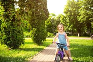 una ragazza con una tuta di jeans cavalca una bici da corsa viola in estate in un parco verde. animazione attiva per bambini, scooter per i più piccoli, bambino felice. copia spazio foto