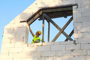 l'operaio edile in cantiere misura la lunghezza dell'apertura della finestra e il muro con un metro a nastro. i cottage sono fatti di blocchi di cemento poroso, indumenti protettivi - elmetto protettivo e un giubbotto foto