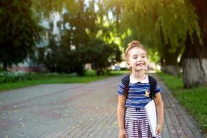 una bambina di aspetto caucasico in uniforme scolastica con uno zaino e il libro e un set di pennarelli. di nuovo a scuola. scuola elementare, sviluppando attività per bambini in età prescolare. spazio per il testo foto