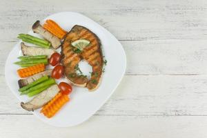 tranci di salmone grigliato fino a cottura, adagiati su un piatto bianco con limone su pesce e verdure, adagiati intorno al piatto su un pavimento di legno bianco. foto