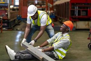 operaio afroamericano che ha un incidente mentre lavora nel sito di produzione mentre il suo collega sta aiutando per la sicurezza sul posto di lavoro e il concetto di emergenza foto