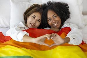 coppia dello stesso sesso matrimonio da razze diverse con bandiera arcobaleno lgbtq per il mese dell'orgoglio mentre si sdraiano insieme sul letto con il gesto del cuore per promuovere l'uguaglianza e le differenze del concetto omosessuale foto