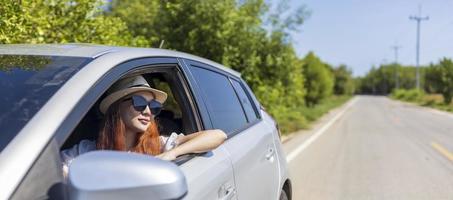 donna con capelli rosso zenzero che indossa occhiali da sole guida auto in un weekend da solista vacanza viaggio guardando fuori dalla finestra in estate con una natura verde lussureggiante in strada di campagna per la libertà e l'avventura foto