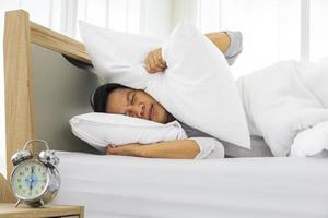 l'uomo si copre pigramente l'orecchio con un cuscino per il forte rumore della sveglia della sveglia al mattino mentre dorme nel suo letto foto