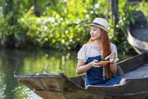 giovane turista asiatico sta viaggiando con una barca di legno nel mercato galleggiante in tailandia e avendo cibo di strada locale per il concetto di turismo del sud-est asiatico foto