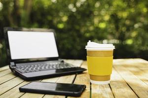 carta della tazza di caffè con un computer portatile e un telefono nel fondo verde della natura. lavorare a distanza o da casa. effetto di messa a fuoco morbida. foto