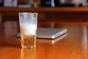 un bicchiere di birra sul tavolo di legno ha lo sfondo del laptop nella caffetteria. birra in tazza con il fondo del taccuino del computer sulla tavola di legno nella barra. bere birra durante il lavoro a casa. foto