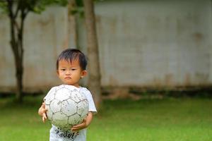 ragazzo asiatico che gioca a calcio al parco. bambino che tiene la palla in mano nel campo in erba. foto