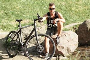 giovane alla moda con una bicicletta seduto su una roccia. t-shirt nera con stampa 23 foto