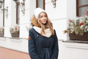 bella ragazza con un cappello lavorato a maglia e una giacca in piedi vicino al muro con decorazioni natalizie. foto