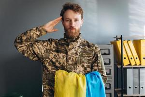 ritratto di un soldato ucraino in uniforme militare che tiene una bandiera gialla e blu e rende omaggio in ufficio foto