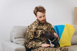 un soldato ucraino in uniforme militare è seduto su un divano con il suo fedele amico, un cane amstaff, sullo sfondo bandiera gialla e blu. foto