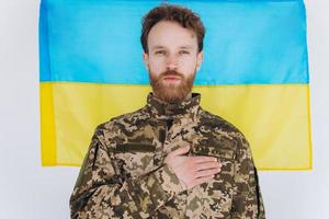 Il soldato patriota ucraino in uniforme militare tiene una mano su un cuore sullo sfondo di una bandiera gialla e blu foto