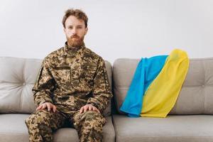 ritratto di un giovane soldato patriota ucraino emotivo in uniforme militare seduto in ufficio su un divano con una bandiera gialla e blu. foto