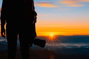 giovane fotografo hipster che tiene la fotocamera con il tramonto sullo sfondo naturale della montagna. foto