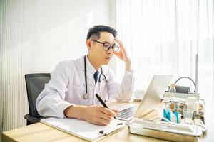 medico asiatico travagliato che lavora al computer alla scrivania nella clinica ospedaliera, preoccupato, pensando a fondo, concetto di assistenza sanitaria per covid19. foto