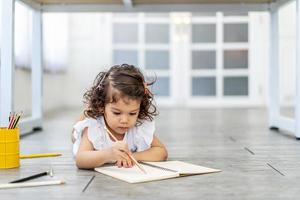 carina bambina che scrive su un notebook sdraiato sul pavimento, imparando il concetto a casa foto