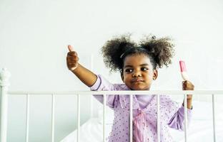 afroamericano carino bambina che tiene mangiare godendo di un delizioso gelato e pollice in alto sul letto in camera da letto foto