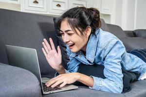 donna asiatica sta riposando sul divano di casa sua, chattando in una videochiamata con un'amica intima sullo schermo del computer portatile, lavorando da casa foto