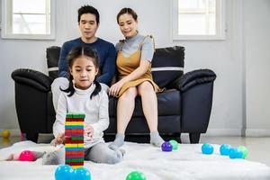 felice famiglia asiatica padre e madre con una piccola ragazza asiatica sorridente che gioca con la torre dell'edificio da blocchi di legno, prendendo le piastrelle a turno da sotto fino a quando non cade, in momenti divertenti a casa foto