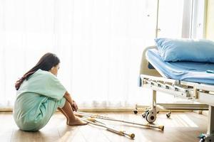 la donna asiatica paziente indossa un abito malato che si sente male e si preoccupa di sedersi e tenere le ginocchia nel pavimento della stanza d'ospedale vicino al letto del paziente, concetto di stile di vita del paziente sanitario e medico. foto