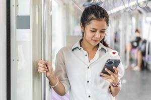 passeggero giovane donna asiatica in piedi e utilizzando il telefono cellulare con l'icona del cuore all'interno della metropolitana vuota o vagone del treno sopraelevato, guarda il messaggio degli amanti, del tempo libero e della vita quotidiana foto