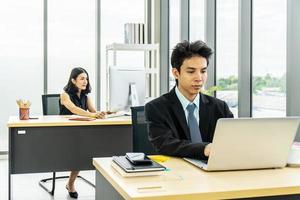 giovane uomo d'affari asiatico seduto alla scrivania che lavora al computer portatile e donna d'affari che lavora al computer portatile nella sua postazione di lavoro in background. foto