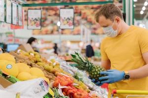 l'immagine orizzontale dell'uomo seleziona l'ananas in negozio indossa una maschera medica e guanti acquista frutta fresca durante l'epidemia di coronavirus o le pose pandemiche di virus nel reparto di alimentari. protezione, prevenzione delle misure foto