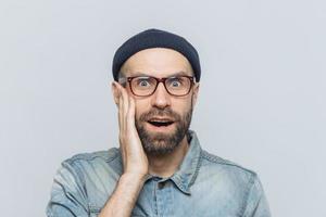 tiro al coperto di un bell'uomo sorpreso fissa la fotocamera, tiene la mano sulla guancia, indossa occhiali e abiti eleganti, isolato su bianco studio backgrpund. persone, espressioni facciali ed emozioni foto