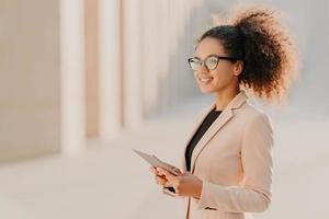 profilo di una donna felice dalla pelle scura con capelli afro, vestita elegantemente tiene in mano il tablet computer passeggiate in strada indossa occhiali ottici collegati a Internet ad alta velocità. stile di vita, tecnologia foto