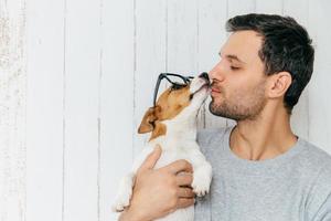 l'inquadratura orizzontale del bel maschio sta lateralmente, bacia il suo cane jack russell terrier, ha un buon rapporto, posa contro uno sfondo di legno bianco con spazio vuoto per la copia. concetto di animali foto