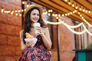 ritratto di donna riposante con capelli scuri, occhi lucenti e labbra ben modellate che indossa cappello e vestito, cena, mangia croissant e beve caffè guardando da parte con un sorriso, avendo buon umore foto